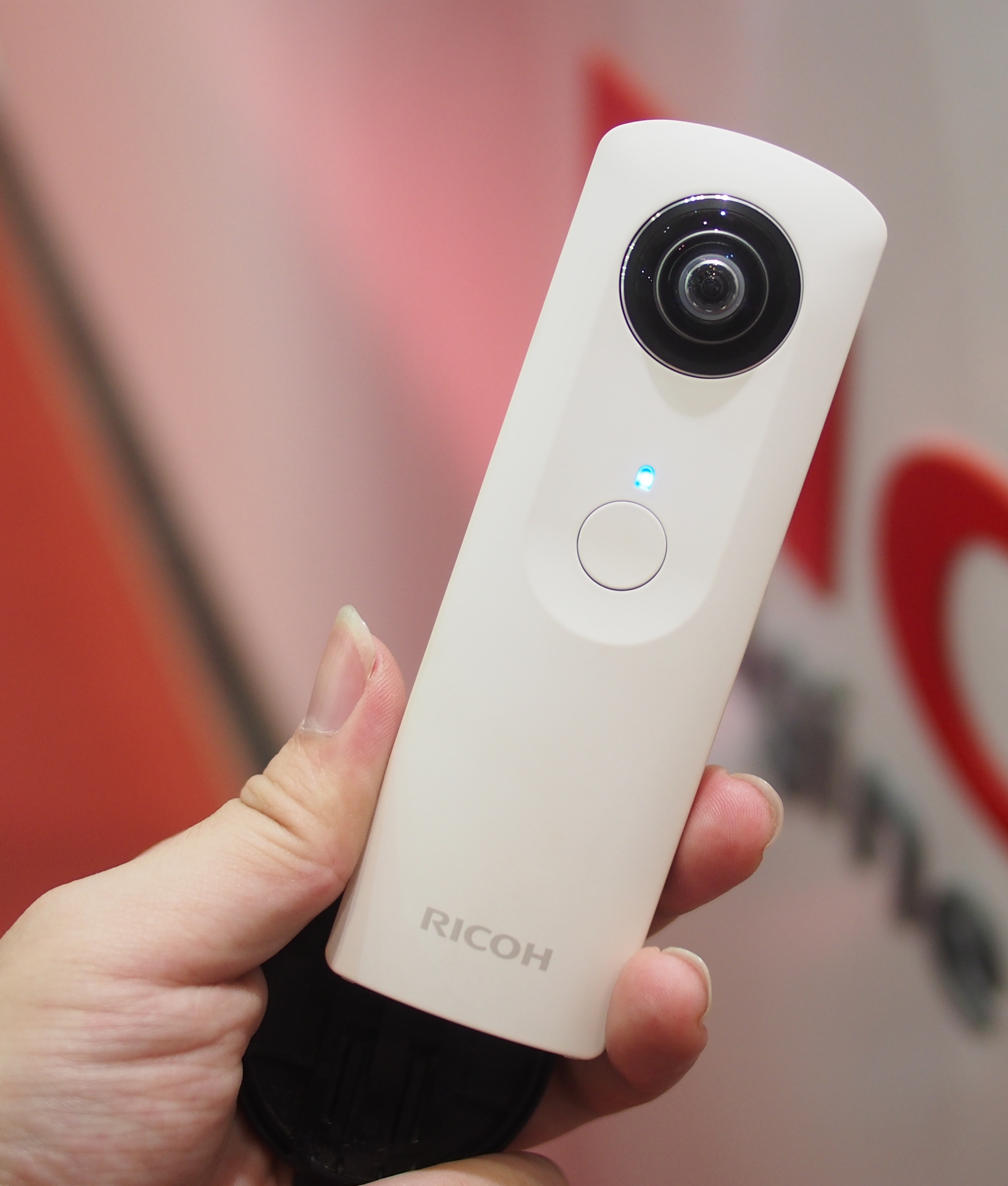 Original white Ricoh Theta held in one hand, showing its fisheye lens and round shutter button