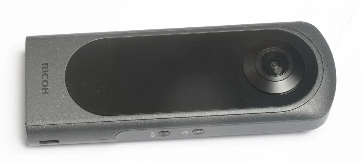 Ricoh Theta X camera on a pale surface with its lens, screen, and side controls visible.